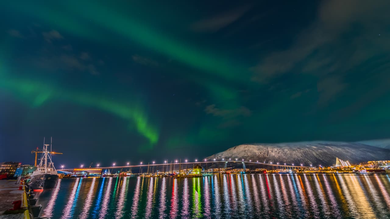 Hermoso Trømso. Aurora boreal sobre un arcoiris creado por el reflejo sobre el agua de las luces del puerto de Trømso, en Noruega.