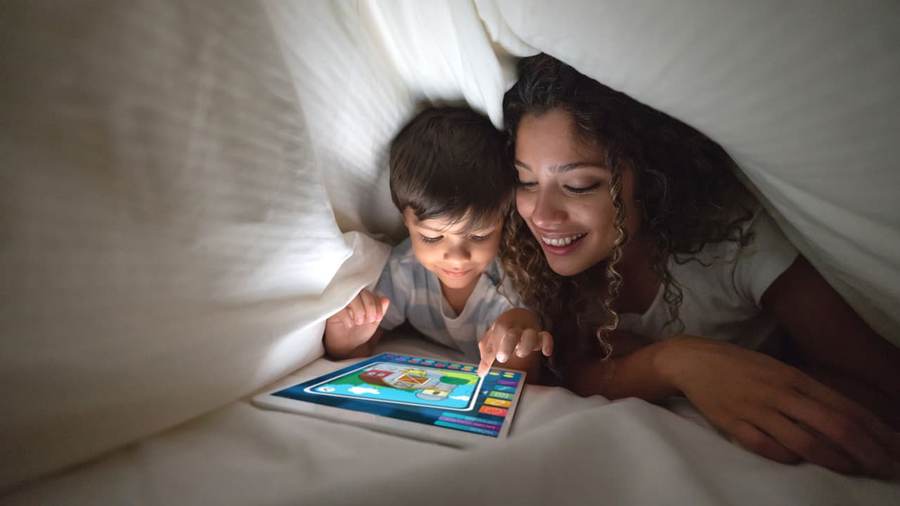 Portrait of a happy mother and son playing on a digital tablet in bed and smiling under the covers