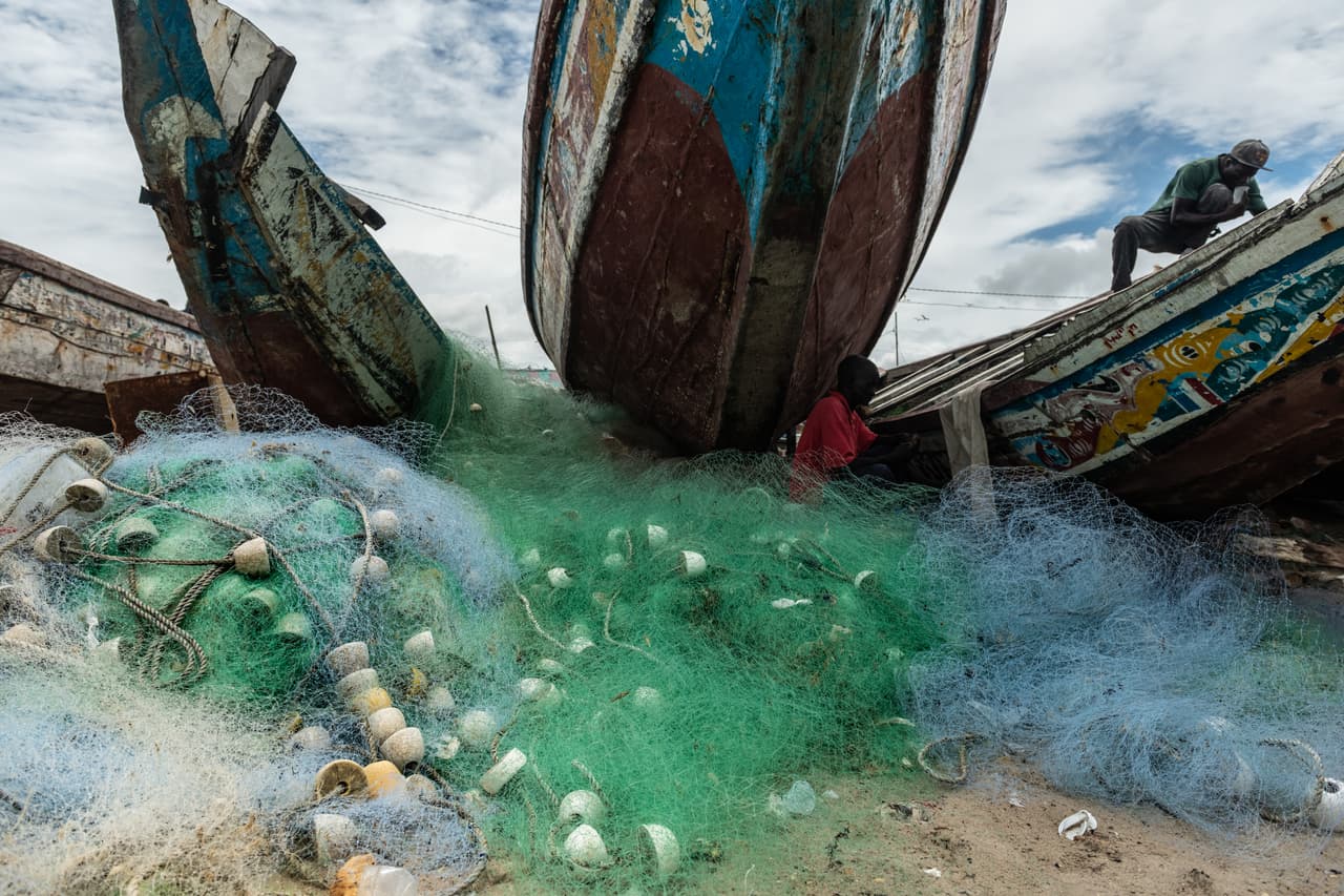 A pile of fishing nets next to Gambian fishermen and their vessels.