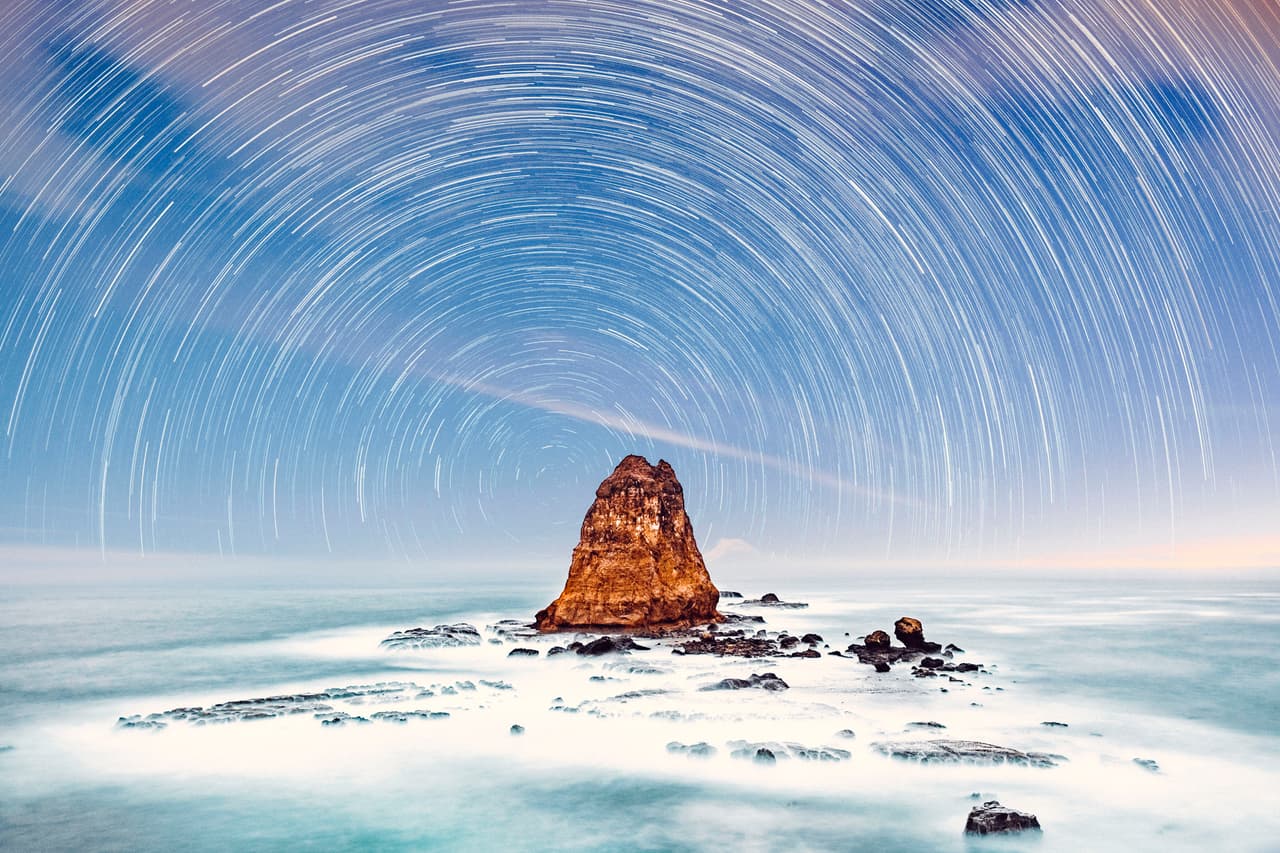 Papuma. Aquí se observan los 'estelas de estrellas' en un cielo un poco nublado formando un arco sobre las rocas y el mar en la playa Papuma en Indonesia.