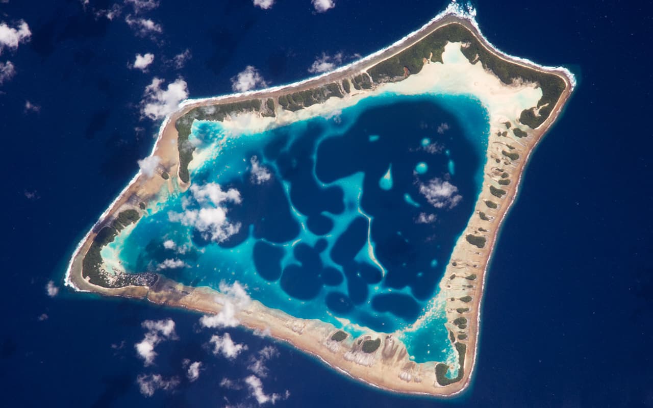 <b>Atolón de Atafu, Tokelau. </b>Con aproximadamente ocho kilómetros de ancho, el atolón de Atafu es el más pequeño de tres atolones que comprende el grupo de islas Tokelau, en el sur del Océano Pacífico. Es actualmente un territorio de Nueva Zelanda aunque se han celebrado referéndums sobre la independencia de las islas. El asentamiento principal en Atafu es un pueblo (casi en el extremo izquierdo, visibles puntos grises) que se ve en esta fotografía de un astronauta. La típica forma de anillo del atolón es el resultado de la acumulación de arrecifes de coral alrededor de una antigua isla volcánica. Con el tiempo geológico, el volcán central se ha hundido
<br>