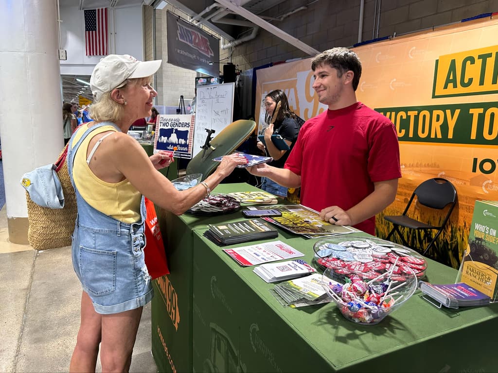 Un miembro del personal de Turning Point Action entrega una bandera estadounidense a un votante en la Feria Estatal de Iowa, en Des Moines, Iowa, el viernes 21 de agosto de 2026. (Foto AP / Hannah Fingerhut)