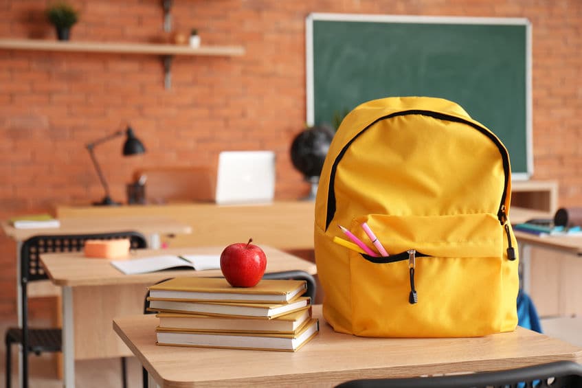 Backpack with fresh apple and books on desk in classroom