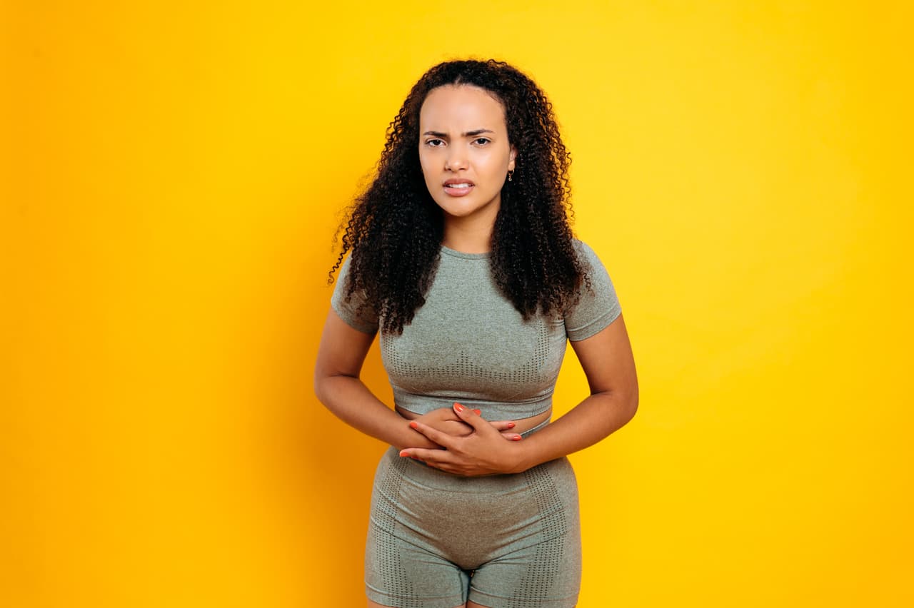 Upset sporty brazilian or hispanic young woman, wearing sportswear, experiencing abdominal pain, spasm, pain during menstruation, holding her hands on a stomach, standing on isolated orange background
