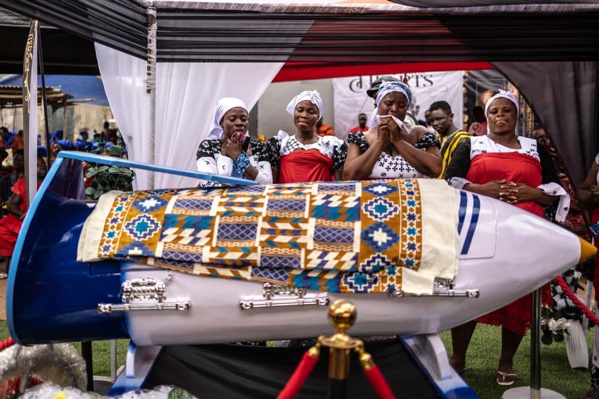 <b>Las lágrimas acompañan al último homenaje:</b> Familiares lloran junto al ataúd con forma de bolígrafo de Samuel Commey Tetteh durante su funeral en Paanor. La peculiar pieza recuerda su trayectoria como profesor y transforma un objeto cotidiano de su profesión en un símbolo de despedida.