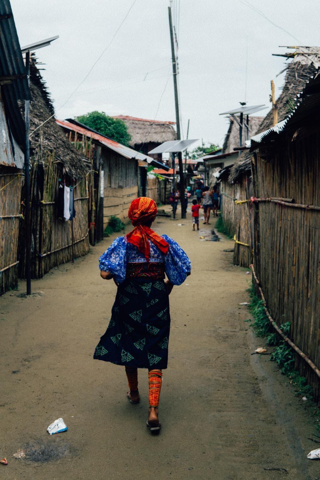A Guna woman in traditional clothing walking along the narrow sandy street on Gardi Sugdub, one of the islands of Guna Yala, Panama. The island is a maze of muddy paths and there are no vehicles, only an elementary school, a police station, a medical clinic, and a few stores with basic supplies and a gas station for the numerous small boats. The rest of the island is filled with housing, some made of palm fronds and others simple, one- or two-story concrete block structures with aluminum roofs and solar panels.